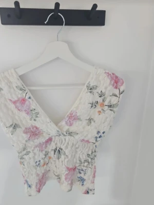 Blommig spets topp med v-ringning - Söt  topp i vit spets med blommigt mönster. Djup v-ringning både fram och bak, ärmlös.