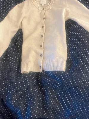 Vit kofta från H&M med knappar - Söt vit kofta från H&M i storlek XS. Koftan har rund halsringning, ribbade muddar och knäppning med bruna knappar framtill. Perfekt att slänga över en topp för en enkel och stilren look.