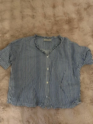 Randig blåvit skjorta med knappar - Kortärmad skjorta med blå och vita ränder och knappar framtill. Skjortan har en avslappnad passform och v-ringad hals. Perfekt för varma dagar och enkel att styla till jeans eller shorts.