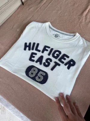 Vit Hilfiger East t-shirt med tryck - Enda defekten är noppor. Vit t-shirt från Hilfiger med blått broderat tryck 'HILFIGER EAST 85' på framsidan. Klassisk rund hals och korta ärmar. Perfekt för en avslappnad och sportig stil.
