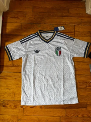 Italien Adidas retro fotbollströja - Snygg retroinspirerad fotbollströja från Adidas med Italiens landslagsmärke. Tröjan är ljusgrå med mörkblå och gula detaljer, V-ringad krage och klassiska Adidas-ränder på axlarna. Tillverkad i lätt och ventilerande material, perfekt för träning eller match.