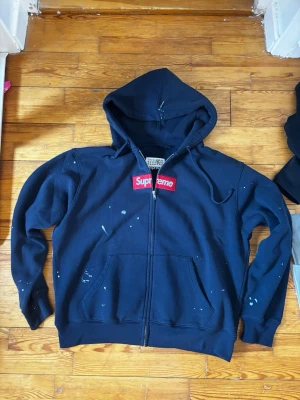 Supreme x Margiela svart hoodie - Svart hoodie med dragkedja från Supreme x Maison Margiela. Klassisk röd Supreme box logo på bröstet och Margiela-etikett i nacken. Hoodien har huva, känguruficka och vita färgstänk som ger en unik look. Perfekt för dig som gillar streetwear och vill sticka ut.