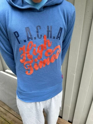 Blå hoodie med tryck P.A.C.H.A - Säljer en blå hoodie med huva och stort tryck på bröstet där det står 'P.A.C.H.A High Times' i marinblått och rött. Hoodien har en klassisk passform och är gjord i mjukt material, perfekt för chill dagar.
