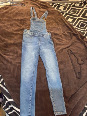 Blå denim hängselbyxor - Snygga blå denim hängselbyxor med klassisk bröstficka och justerbara axelband. Byxorna har smal passform, fram- och bakfickor samt metallspännen. Perfekta för en avslappnad och trendig look. Materialet är slitstarkt bomullsdenim.