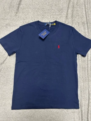 Marinblå t-shirt från Polo Ralph Lauren - Klassisk marinblå t-shirt från Polo Ralph Lauren med rund halsringning och den ikoniska röda logobrodyrn på bröstet. Tillverkad i mjuk bomull och har en normal passform. Perfekt basplagg för en clean och stilren look.