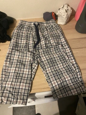 Rutiga pyjamasbyxor med dragsko - Ett par klassiska pyjamasbyxor i mjuk bomull med rutigt mönster i blått, vitt och rött. Byxorna har resår i midjan och en mörkblå dragsko för justerbar passform samt två stora fickor framtill. Perfekta för mysiga hemmakvällar.