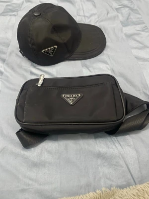 Svart keps från Prada i nylon - Snygg svart keps från Prada i glansigt nylonmaterial med klassisk böjd skärm. Kepsen har Pradas ikoniska trekantslogga i metall framtill och justerbar rem baktill för perfekt passform.