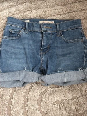 Levi's blå jeansshorts med uppvik - Säljer ett par klassiska blå jeansshorts från Levi's med uppvikta benslut och femficksmodell. Shortsen har normal passform, bälteshällor och dragkedja med knapp. Perfekta för varma dagar och enkel att matcha med allt.