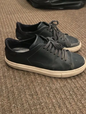 Axel Arigato skor - Stilrena mörkblåa Axel Arigato skor med lågt skaft och vita sulor. Skorna har klassisk snörning och är tillverkade i slätt material som ger en clean look. Passar perfekt till jeans eller chinos för en avslappnad stil.