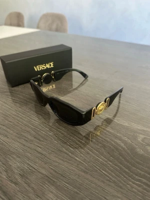 Svarta solglasögon från Versace - Snygga svarta solglasögon från Versace med rektangulära bågar och guldfärgade detaljer på sidorna, inklusive den ikoniska Medusa-loggan. Tillverkade i Italien och levereras med originaletui. Perfekt accessoar för en trendig look.