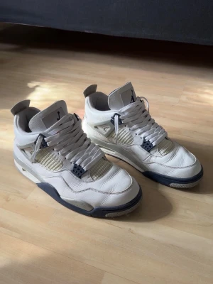 Jordan 4 - Säljer ett par vita Jordan 4 sneakers med meshpaneler och detaljer i mörkblått. Skorna har klassisk siluett, tjock sula och ikoniska plastdetaljer vid snörningen. Perfekta för dig som gillar streetwear och vill sticka ut med en tidlös modell. Tyvärr saknar skorna sula och har en liten skada som syns på sista bilden. Hör av dig ifall du har fler frågor! 