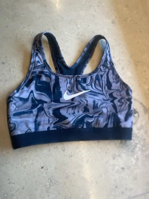 Blå sport-bh med mönster från Nike - Säljer en sport-bh från Nike i marinblå och grå toner med ett abstrakt mönster. Bh:n har brottarrygg och ett vitt Nike-märke framtill. Perfekt för träning och ger bra stöd. Materialet känns som syntet och polyester.
