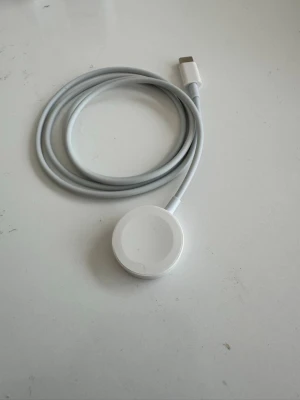 Apple Watch laddkabel USB-C - Säljer en vit magnetisk laddkabel med USB-C kontakt, perfekt för smartklockor och wearables. Kabeln är rund och flexibel med en slät yta och laddplattan är i plast. Enkel att använda för trådlös laddning av kompatibla enheter.