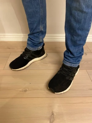 Svarta sneakers med vit sula - Snygga svarta sneakers med stickad ovandel och vit, dämpad sula. Skorna har svarta snören och plastdetaljer på sidorna. Perfekta för dig som gillar sportig och stilren look. Passar till jeans eller träningskläder.