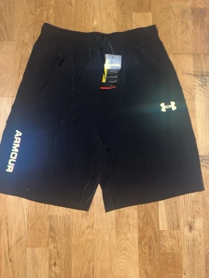 Svarta träningsshorts från Under Armour - Snygga svarta träningsshorts från Under Armour med logga och text på vänster ben. Shortsen har elastisk midja med snörning och reflekterande detaljer baktill. Perfekta för gymmet eller löprundan.