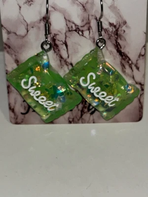 Gröna örhängen med texten Sweet - Unika örhängen i form av gröna godisförpackningar med holografiska detaljer och texten 'Sweet' i vitt. Tillverkade i plast med silvriga krokar. Perfekta för dig som gillar färgglada och lekfulla accessoarer. Örhängens krokarna är i rostfritt stål 
