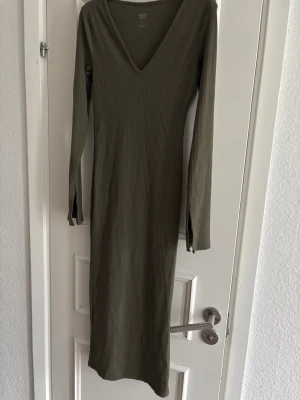 Olivgrön ribbad långklänning - Snygg olivgrön långklänning med v-ringning och långärmad design. Klänningen är ribbad och har en kroppsnära passform som framhäver siluetten. Perfekt för dig som gillar stilrena och enkla plagg med en modern touch.