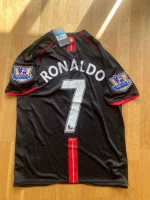 Manchester United Ronaldo tröja svart - Snygg svart Manchester United matchtröja med Ronaldo och nummer 7 på ryggen. Tröjan har röda detaljer, Premier League-patchar på ärmarna och AIG som sponsor på bröstet. Tillverkad i lätt och ventilerande material, perfekt för fotboll eller streetwear.