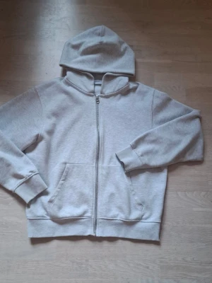 Ljusgrå zip hoodie med huva - Säljer en stilren ljusgrå hoodie med dragkedja framtill och stor ficka på magen. Hoodien har ribbade muddar vid ärmslut och nederkant samt en klassisk huva. Perfekt för en avslappnad och chill look.