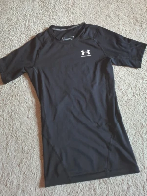 Svart träningströja Under Armour - Säljer en svart, tight och kortärmad träningströja från Under Armour. Tröjan har rund halsringning och är tillverkad i ett stretchigt funktionsmaterial som andas. Perfekt för gymmet eller löpning. Diskret logga på bröstet och nacken.