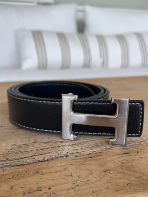 Herme’s bälte  - Snyggt svart Herme’s skärp i skinn med ett stort, silverfärgat H-format spänne. Bältet har vita kontrastsömmar längs kanterna som ger en stilren touch. Perfekt accessoar för att lyfta vilken outfit som helst. Sprillans ny aldrig använd.