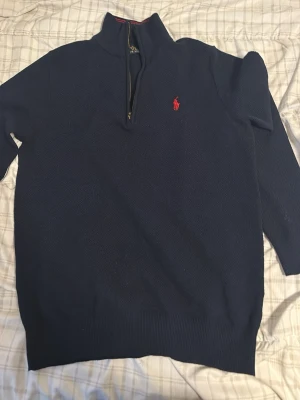Mörkblå stickad tröja Polo Ralph Lauren - Mörkblå stickad tröja från Polo Ralph Lauren med halv dragkedja och röd broderad logga på bröstet. Tröjan har hög krage med röd detalj och ribbade muddar vid ärmslut och nederkant. Perfekt för lager på lager och klassisk stil. ( använd ett par få gånger ) 