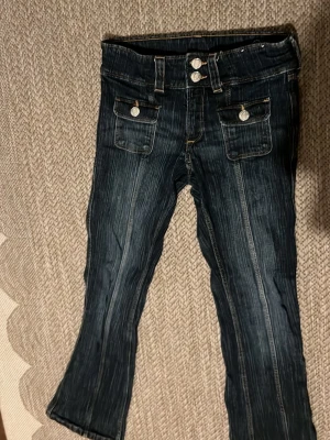 Mörkblå bootcut jeans med fickor - Säljer ett par mörkblå bootcut jeans med dubbla knappar i midjan och framfickor med lock och silverfärgade knappar. Jeansen har markerade sömmar och en lätt tvättad look. Perfekta för dig som gillar en retrostil och vill ha något unikt i garderoben.
