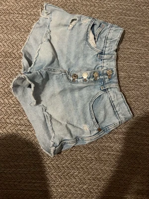 Ljusa jeansshorts med slitningar - Säljer ett par ljusblå jeansshorts med råa kanter och slitna detaljer. Shortsen har knappgylf med fyra synliga knappar och klassiska fem fickor. Perfekta för sommaren och ger en avslappnad streetstil.