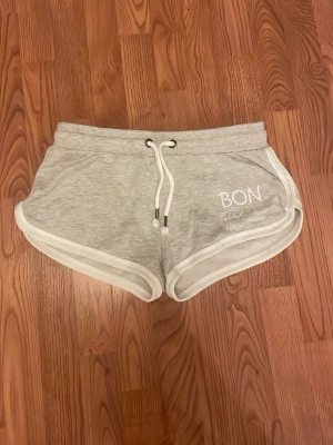Grå mjukisshorts med snörning - Snygga ljusgrå mjukisshorts med vita kanter och snörning i midjan. Tryck med texten 'bondelid' på ena sidan. Perfekta för sommarens chill eller träning. Sköna och lätta att matcha med en enkel t-shirt eller hoodie.
