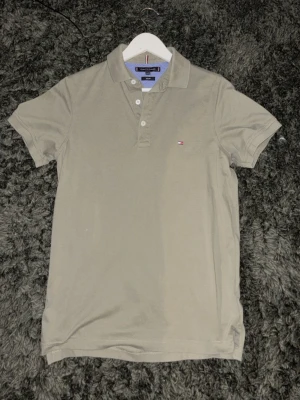 Beige pikétröja från Tommy Hilfiger - Klassisk beige pikétröja från Tommy Hilfiger med liten logga på bröstet. Tröjan har krage, knappar framtill och korta ärmar. Perfekt till jeans eller shorts för en clean och enkel stil.