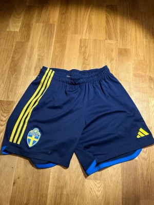 Blå Sverige shorts med gula detaljer - Säljer ett par mörkblå sportshorts med gula ränder och Sveriges landslagsemblem på vänster ben. Shortsen har elastisk midja och blå kant längst ner. Perfekta för fotboll eller träning.