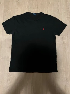 Svart t-shirt med v-ringning - Svart t-shirt med v-ringad hals och korta ärmar. Liten röd logga broderad på bröstet. Tillverkad i mjuk bomull som känns skön mot huden. Enkel och stilren design som passar till det mesta.