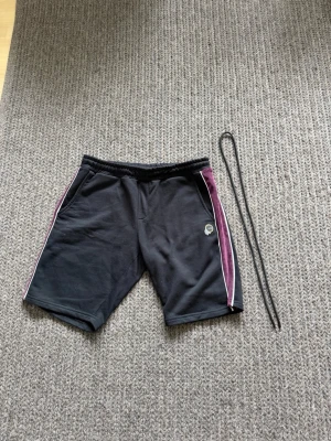 Svarta shorts - Snygga svarta shorts från Le VICHY med lila och vita ränder längs sidorna. Shortsen har resår i midjan och medföljande snöre för justering. Tillverkade i mjukt material, perfekta för en avslappnad stil.