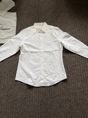 Vit klassisk skjorta i linne - Stilren vit skjorta med klassisk krage och knappar framtill. Skjortan har lång ärm och är tillverkad i linne, perfekt för en clean och enkel look. Passformen är normal och tyget känns lätt och bekvämt.