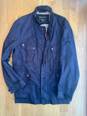 Marinblå jacka från Woolrich - Snygg marinblå utilityjacka från Woolrich med fyra framfickor med lock och knappar, hög krage och dragkedja samt knappstängning. Tillverkad i ett slitstarkt bomullsmaterial och har en klassisk, rak passform. Perfekt för dig som gillar stilren och funktionell design.