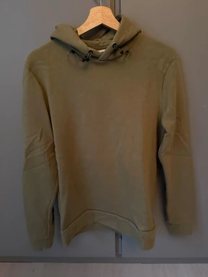 Grön hoodie med tryck - Säljer en grön hoodie med huva och dragsko. På ryggen står det 'UTILITY' i stort tryck och på ärmarna finns texten 'DIVERSE' och 'ATTIRE' i mörkgrönt. Hoodien har en avslappnad passform och är gjord i mjukt material, perfekt för en chill och street-inspirerad look.