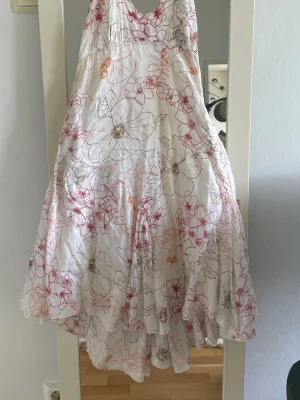 Vit långklänning med blommigt mönster - Luftig och somrig vit långklänning med tunna axelband och blommigt mönster i rosa, rött och beige. Klänningen har en v-ringad hals och ett svepande fall som ger en romantisk känsla. En lång sido dragskedja , Perfekt för varma dagar och midsommarfirande.