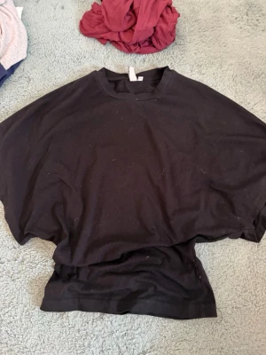 Svart loose topp med korta ärmar - En svart topp med loose passform och korta ärmar. Toppen har rund halsringning och är tillverkad i ett mjukt bomullsliknande material. Perfekt för en avslappnad och enkel stil.