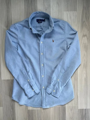 Ljusblå manchesterskjorta från Ralph Lauren - Säljer en ljusblå manchesterskjorta från Ralph Lauren med klassisk krage och knappar framtill. Skjortan har lång ärm och den ikoniska broderade loggan på bröstet. Perfekt för dig som gillar stilrena och tidlösa plagg.