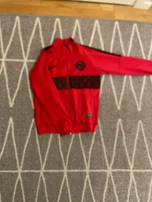 Röd PSG träningsjacka från Nike - Snygg röd träningsjacka från Nike med Paris Saint-Germain-logga på bröstet och svart mönstrad rand över magen. Jackan har hel dragkedja, ståkrage och muddar vid ärmslut och midja. Perfekt för fotbollsträning eller streetwear.