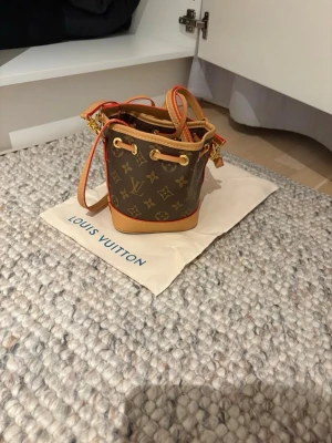 Louis Vuitton monogram bucket bag - Snygg liten bucket bag från Louis Vuitton i klassiskt brunt monogrammönster med beige och ljusbruna detaljer. Väskan har justerbar axelrem och guldfärgade metalldetaljer. Perfekt för dig som vill ha en ikonisk och trendig accessoar.