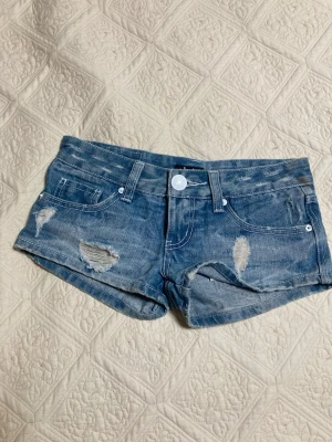 Blå slitna jeansshorts -  Säljer ett par blå jeansshorts med slitna detaljer och råa kanter. Shortsen har låg midja, fem fickor och vita knappar på bakfickorna. Perfekta för sommaren eller i höst med strumpbyxor. 38cm rakt över, pris diskuteras, de är köpta i en vintage butik i Sydkorea. OBS jag kan snarast leverera 21/7 för att jag är på resa. Samt finns det små fläckar