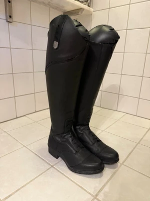 	Ridstövlar Veganza Svart 40 Regular/Wide - Snygga svarta ridstövlar i läder med högt skaft och dragkedja baktill. Klassisk design med snörning framtill och diskret metallmärke vid sidan. Perfekta för dig som vill ha stilrena och funktionella stövlar till ridning. Jag köpte dom för 1439 kr. Mitt pris är 850