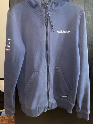 Blå zip-hoodie från Race Marine - Mörkblå hoodie med dragkedja från Race Marine. Har huva med snören, två fickor fram och tryck på bröstet samt ärmen. Tillverkad i mjukt sweatshirtmaterial, perfekt för chill dagar. Klassisk passform och ribbade muddar. Är lite sliten men fortfarande i fint skick.