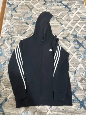 Svart hoodie med vita ränder  (ADIDAS) - Svart hoodie med dragkedja och huva. Klassiska vita ränder längs ärmarna och över ryggen. Mjuk och bekväm passform, perfekt för en sportig och avslappnad stil. Materialet känns som bomullsblandning.