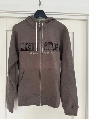 Brun zip-hoodie från Lexington - Brun hoodie med dragkedja från Lexington. Stor Lexington-logga i marinblått framtill, vita snören i huvan och två fickor. Klassisk passform och mjukt material, perfekt för chill dagar. Tjocka muddar vid ärmslut och nederkant.