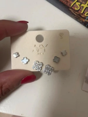 Söta blommiga örhängen i silver - Två par små örhängen i form av blommor. Ett par är silverfärgade med glittrande stenar och det andra paret har en ljus pärlemorfärgad yta. Perfekta för att ge en söt touch till din stil.