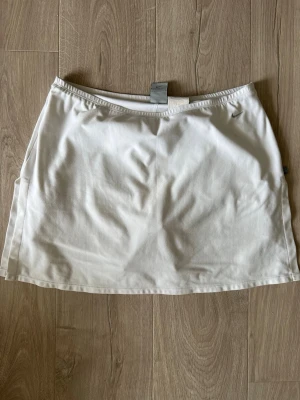 Vit sportig kortkjol från Nike - Snygg vit kortkjol från Nike med diskret logga framtill. Kjolen är tillverkad i ett lätt och funktionellt material som andas, perfekt för sportiga aktiviteter som tennis. Elastisk midja för skön passform.