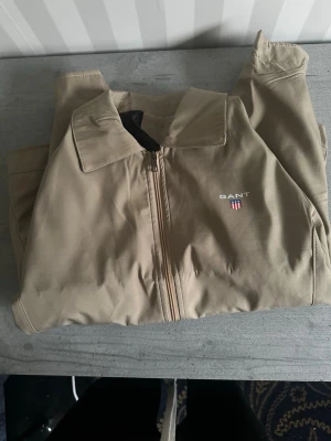 Beige jacka från GANT med dragkedja - Snygg beige jacka från GANT med klassisk krage och dragkedja framtill. Jackan har två sidofickor och diskret GANT-logga med sköld på bröstet. Perfekt för dig som gillar stilrena och tidlösa plagg.