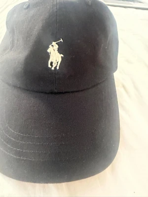 Svart keps från Polo Ralph Lauren - Säljer en svart keps från Polo Ralph Lauren med den klassiska broderade polospelaren framtill. Kepsen har böjd skärm och justerbar rem bak. Tillverkad i bomull för en bekväm och snygg look. Perfekt accessoar för en sportig och stilren outfit.